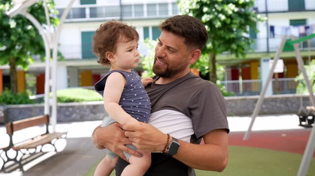 Loving father holding his toddler son in his arms while pointing at something in a public park, sharing a happy and tender moment of complicity and family bonding on a sunny day