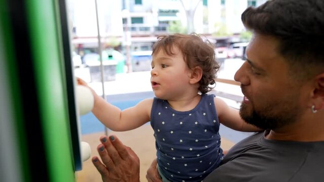 Smiling father holding his cute toddler son while the child happily plays with a colorful abacus in a public park, showcasing a moment of joyful family bonding and early development