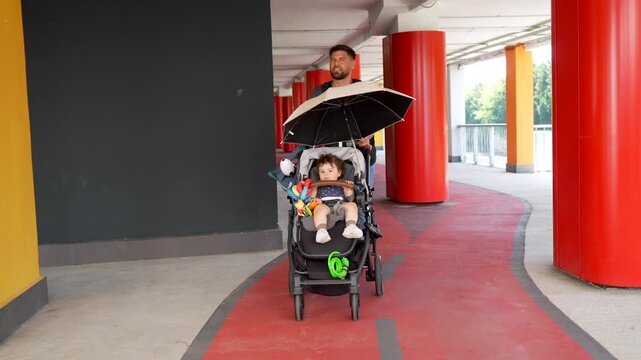 Young father walking his little son in a stroller along a pedestrian path with colorful pillars, protecting him from the sun with a small umbrella attached to the pram