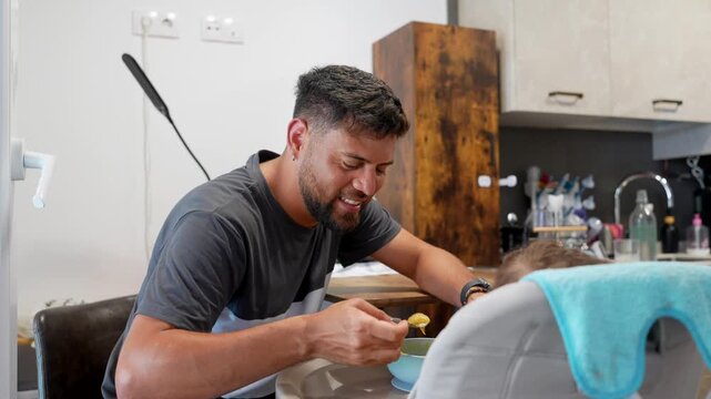 Loving young father with a beard smiling while feeding his adorable baby boy with a spoon, sitting together at the kitchen table. The infant is in a high chair eating puree