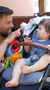 Loving father with a beard smiling and playing with his adorable baby boy sitting in a stroller, showing him a colorful toy and creating a strong bond between parent and child outdoors