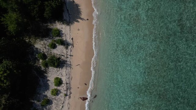 Top down aerial spinning descent onto tropical beach with clear turquoise water and woman walking towards gentle waves in 4k