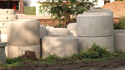 Zdjęcie przedstawia składowisko prefabrykowanych kręgów betonowych na placu budowy. Szare elementy przeznaczone do budowy studzienek kanalizacyjnych stoją na ziemi w otoczeniu roślinności. © tomo81