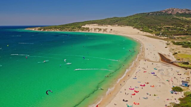 Cinematic aerial footage of Valdevaqueros Beach in Tarifa, Spain, featuring turquoise waters, white sand, coastal dunes and kitesurfers enjoying strong winds along the Atlantic coastline.