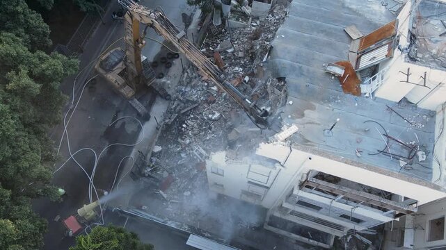 Aerial demolition with excavator removing building facade during urban renewal project, drone orbits as crane pushes and rips ceiling