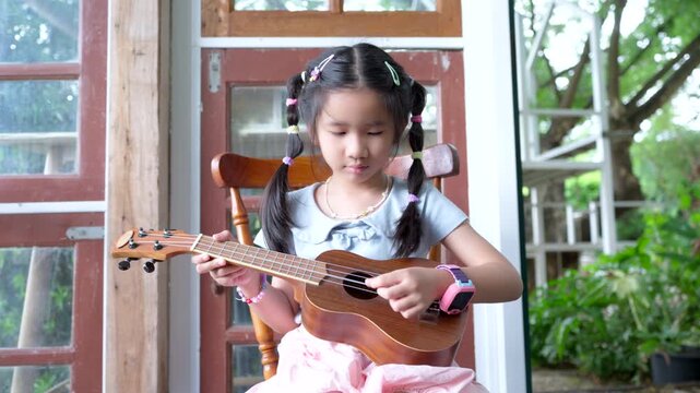 Cute little Asian girl playing ukulele happily while sitting on a wooden chair, childhood learning and music education concept.