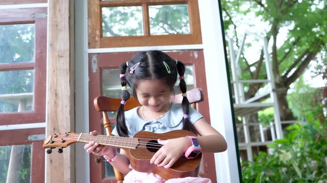 Cute little Asian girl playing ukulele happily while sitting on a wooden chair, childhood learning and music education concept.