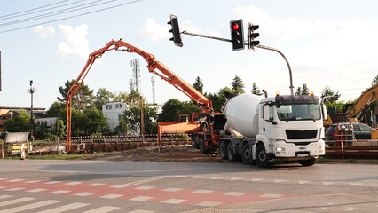 Zdjęcie przedstawia miejski plac budowy na skrzyżowaniu z pracującą betoniarką oraz rozłożoną pompą do betonu. Nad pojazdami widoczna jest sygnalizacja świetlna oraz wysokie ramię maszyny. © tomo81