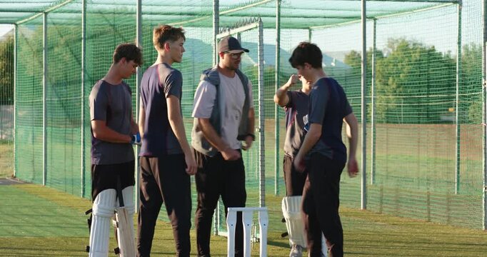 Diverse male coach clapping, urging teenage boys in nets, huddling, batsman rubbing cricket ball
