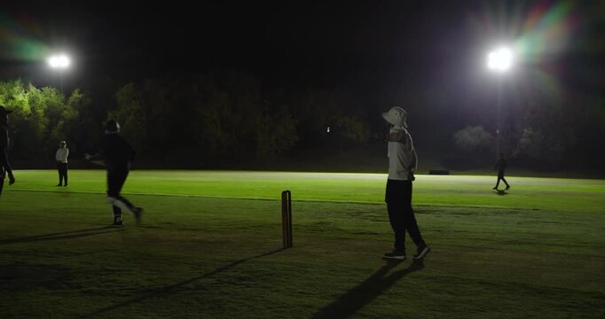 Group of cricket players reacting to bowler run on pitch by tracking red ball, floodlights