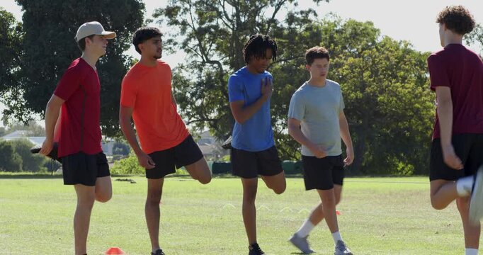 Diverse male friends warming up before drill, stretching on park field marked by orange cones