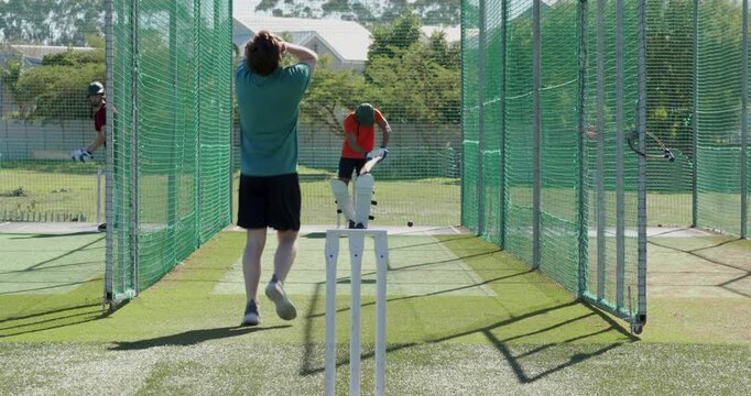 Male bowler training in cricket net signaling running and bowling red ball, batsmen rotating