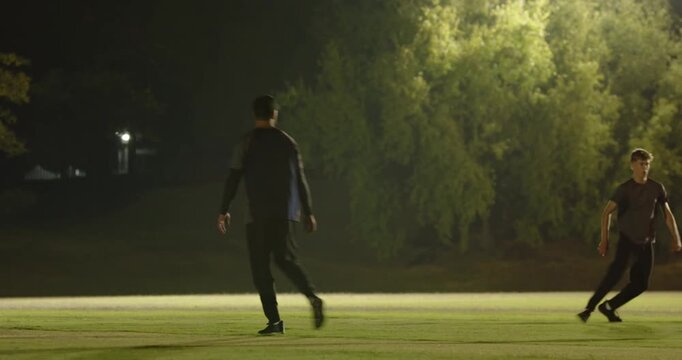 Male bowler launching deliveries twice under floodlights, batsman swinging cricket bat missing ball