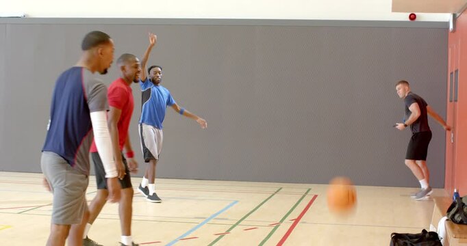 Diverse male group passing basketball in gym, calling coach scrolling tablet, huddling
