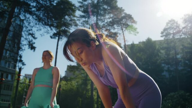 Woman exhales with fatigue after completing a jump rope workout while her twin sister continues exercising with dumbbells nearby, highlighting endurance and strength training