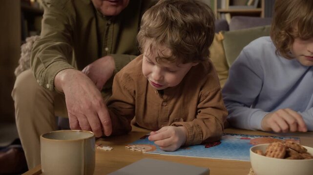 Zoom out shot of senior Caucasian grandfather helping young children assemble jigsaw puzzle at home. Clip is ideal for family bonding, intergenerational connection, and retirement lifestyle ads