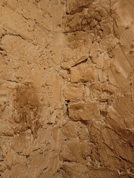 Close-up textured photograph of an old interior masonry brick wall corner painted in a warm sandy beige. This high-quality rustic backdrop highlights heavily weathered limestone blocks and rough cemen