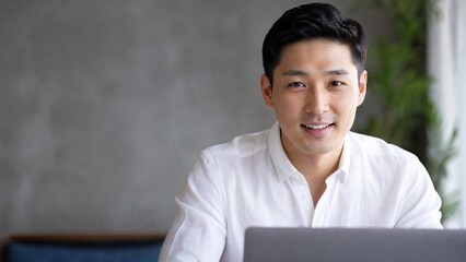 Obraz premium Generative AI, Smiling Asian businessman looking at camera while working on laptop at home office
