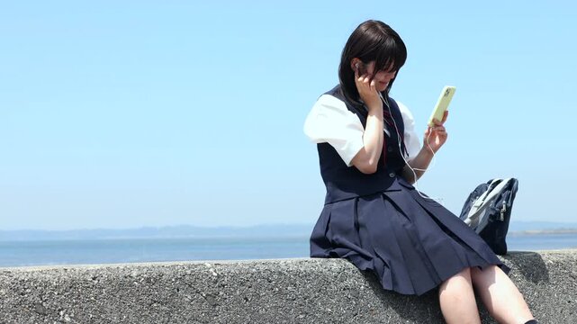 海辺の堤防に座りスマートフォンで音楽を楽しむ女子高校生