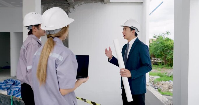 Asian male manager gestures during slow motion site discussion while female engineer uses laptop, guiding construction document review process and engineering supervision inside unfinished building.
