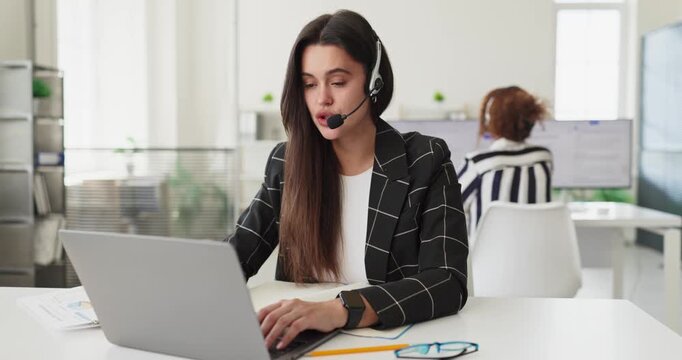 Operator woman with headset microphone on laptop. In a bright office she provides support during a call. Modern helpdesk setting and bright workspace. Clear concise customer service concept.