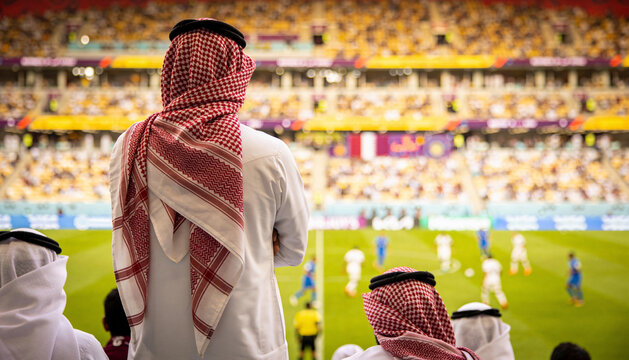 Watching man wearing thobe and shemagh following match on pitch with players and referee at stadium