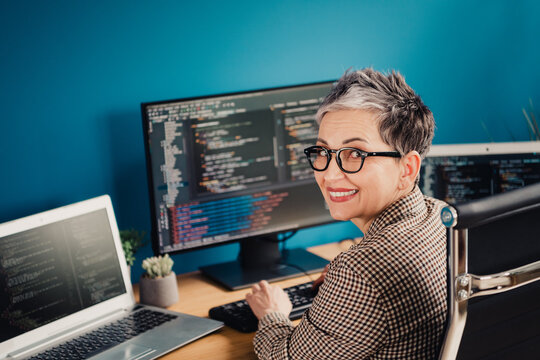 Smiling female programmer at desk coding on multiple monitors in a modern home office working on software development and web coding
