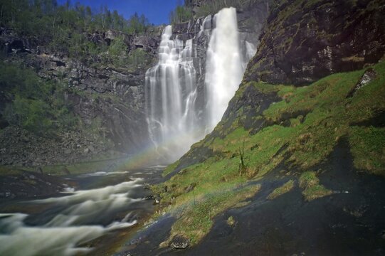 Picturesque waterfall cascading over rocks into a river, surrounded by lush greenery and forest in the background, Regenborgen, Skjervefossen, Voss, Norway
