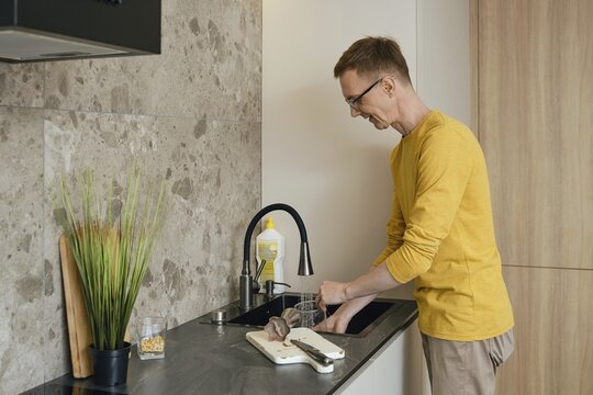A man in a yellow long-sleeve shirt is washing utensils at a sleek kitchen sink. Natural light highlights the modern decor, while the kitchen is organized and well-kept, perfect for food preparation