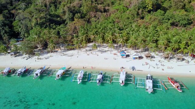Orbit shot over row of outrigger boats moored along pristine white sand beach with sunbathers, beach huts and coconut palm jungle backdrop. El Nido, Philippines