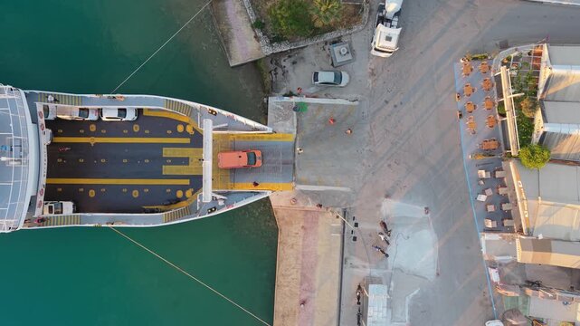 Top down drone view of a vehicle ferry docked at a harbor terminal. Cars and trucks board the vessel via loading ramp beside waterfront buildings and clear coastal waters.