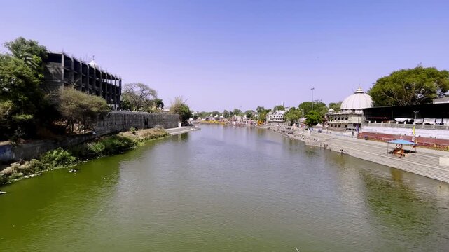 Handheld shot from Narasingh Ghat Bridge overlooking Shri Ram Ghat and the sacred Kshipra River in Ujjain. Temple ghats, riverfront architecture, and pilgrimage surroundings visible during daylight.