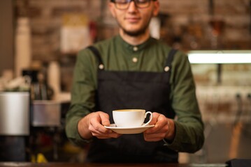 A smiling male barista holding and serving a white cup of hot coffee