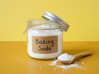 Glass jar of baking soda with a wooden spoon on a yellow and brown surface © Dean