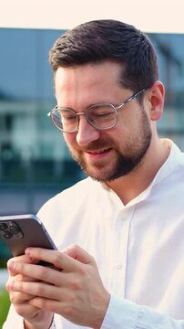 Bearded businessman in a white shirt holding a smartphone, typing messages outdoors, successful entrepreneur or manager working remotely. Slow motion