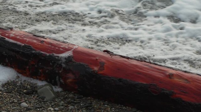 Driftwood waves beach, white ocean foam washes over a red wooden log on a rough, wet pebble shoreline