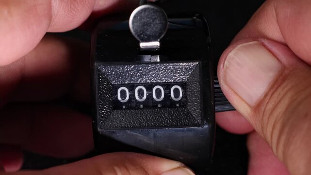 Close-up of a mechanical tally counter displaying clicks. Hand is holding the device, highlighting the worn black plastic casing and textured counter dial.
