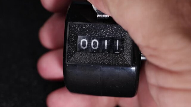 Close-up view of a handheld manual tally counter displaying numbers as it is pressed, showing the counting process from to on a black background.