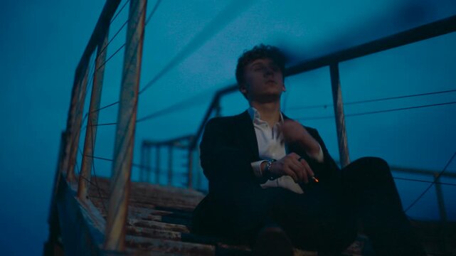 Thoughtful young businessman in a suit smoking a cigarette alone on outdoor stairs at twilight