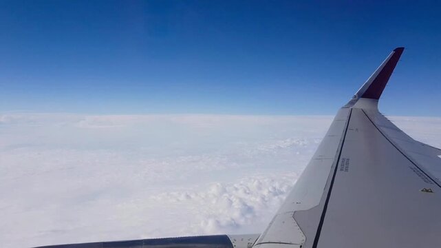 View from a passenger aircraft window showing the wing extending above a thick layer of white clouds during cruise flight. Clear blue sky and cloudscape create a peaceful aviation travel scene at high