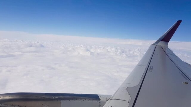 View from a passenger aircraft window showing the wing extending above a thick layer of white clouds during cruise flight. Clear blue sky and cloudscape create a peaceful aviation travel scene at high
