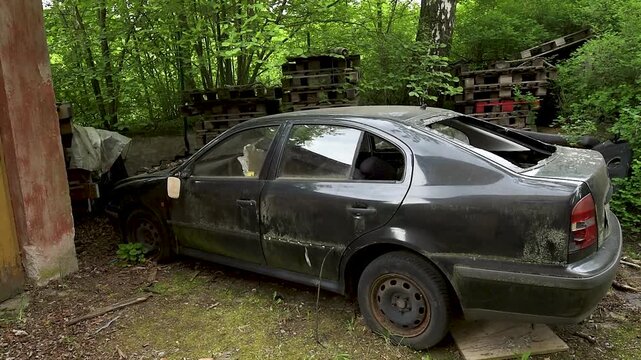A side profile view of a neglected, dirty black sedan abandoned outdoors next to a building and stacked wooden pallets. The car shows signs of decay, covered in moss and grime, with its rear window