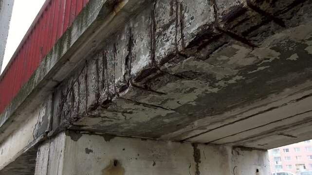A close-up view underneath a badly damaged concrete bridge structure showing severe erosion and spalling. The crumbling concrete has fallen away, exposing heavily rusted steel reinforcement rebar grid