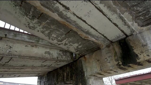 A close-up view underneath a badly damaged concrete bridge structure showing severe erosion and spalling. The crumbling concrete has fallen away, exposing heavily rusted steel reinforcement rebar grid