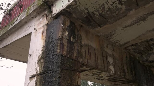 A close-up view underneath a badly damaged concrete bridge structure showing severe erosion and spalling. The crumbling concrete has fallen away, exposing heavily rusted steel reinforcement rebar grid