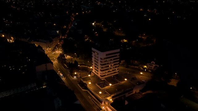 An aerial night view captures a dimly lit street slicing through a city center, with a prominent multi-story modern administrative high-rise building illuminated on the right. Warm streetlights scatte