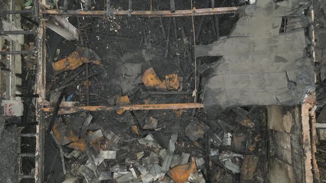 An aerial close-up view reveals the charred metal and wooden skeletal frame of a building devastated by fire. The structure shows severe damage with burnt beams, ash, and collapsed roofing components.