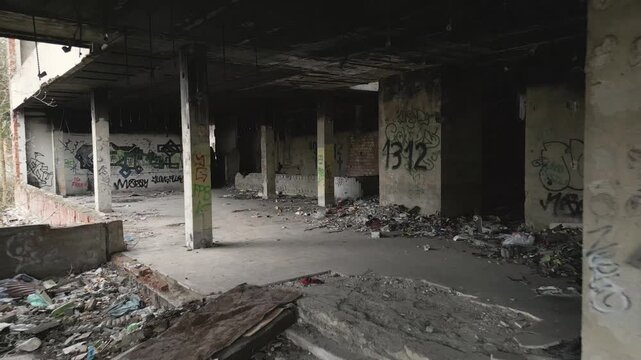 The interior of a decaying, abandoned concrete building is filled with debris, scattered garbage, and walls covered in colorful graffiti tags. The stark, empty industrial shell features rough supporti