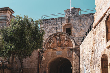 Obraz premium view from behind mdina gate, malta with limestone brick and detailed statues above the archway with clear blue skies overhead in summer sunlight and green lush tree to the left