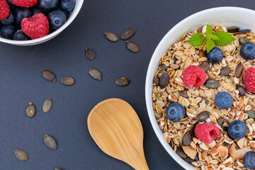 Healthy breakfast muesli bowl with fresh raspberries blueberries and pumpkin seeds, top view of organic granola porridge with wooden spoon on dark background. © JaRu Creative Studio